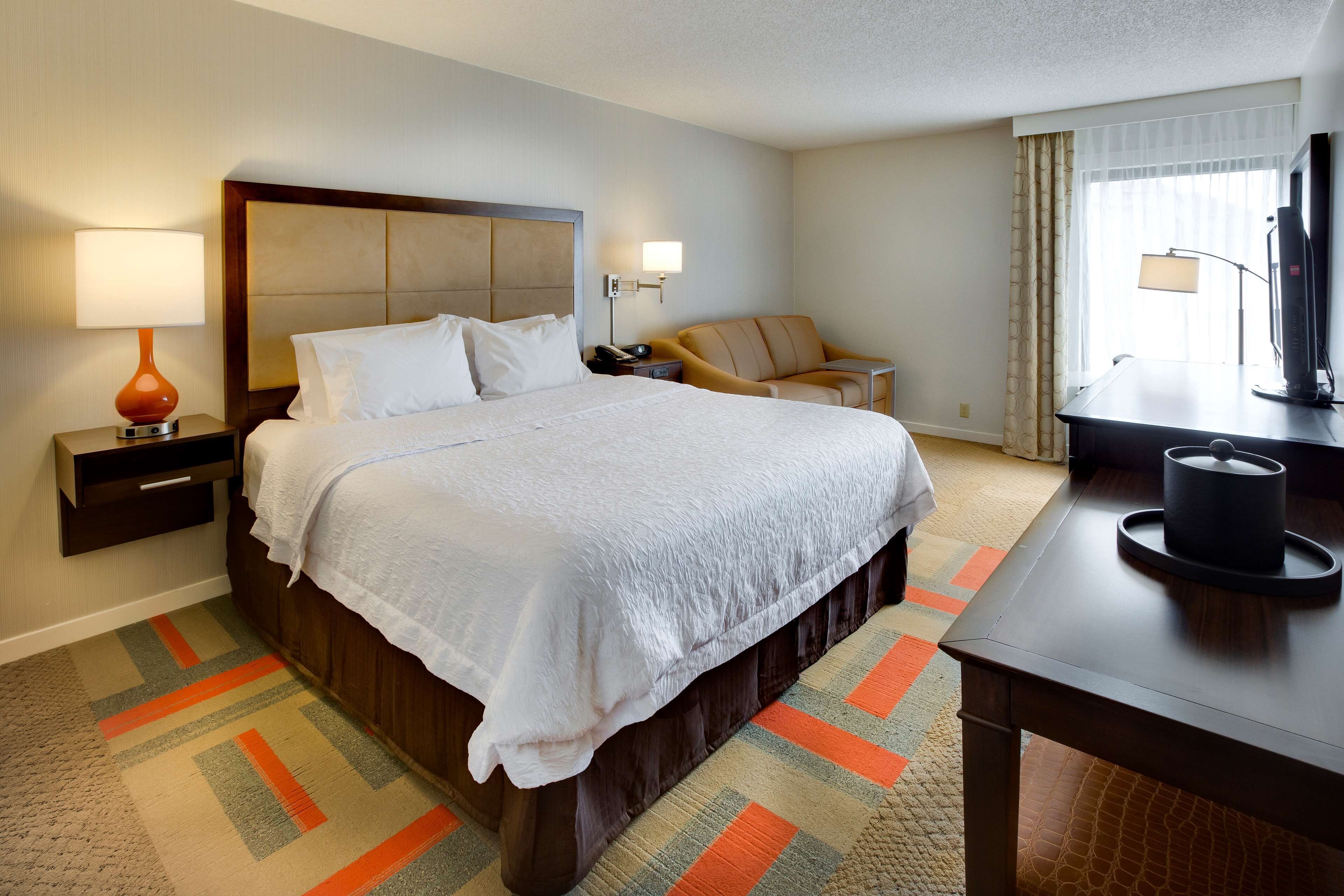 A hotel room with a large bed, a desk, a chair, a TV, and a lamp.