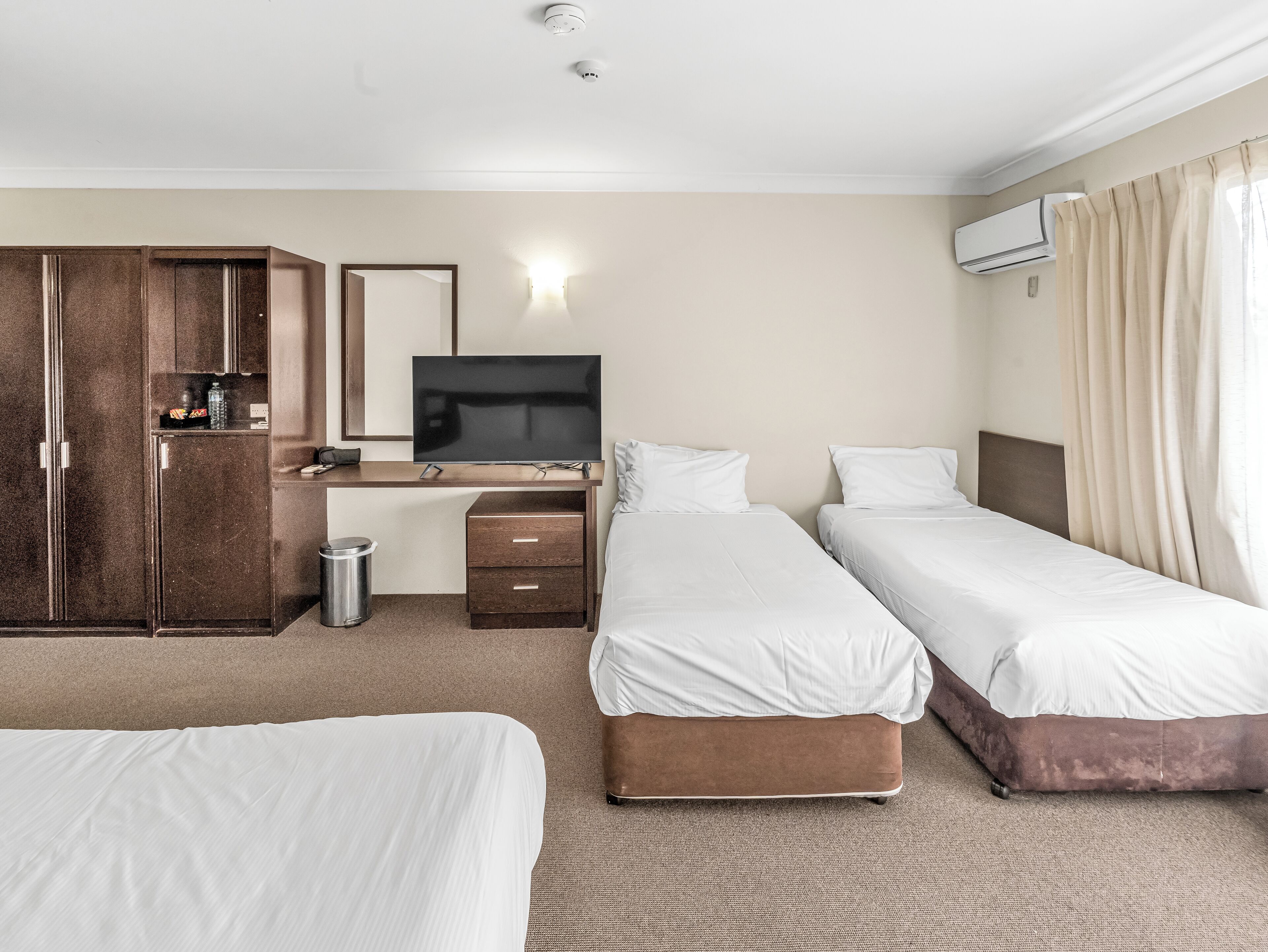 A hotel room with two beds, a desk with a TV, a wardrobe, and an air conditioning unit.