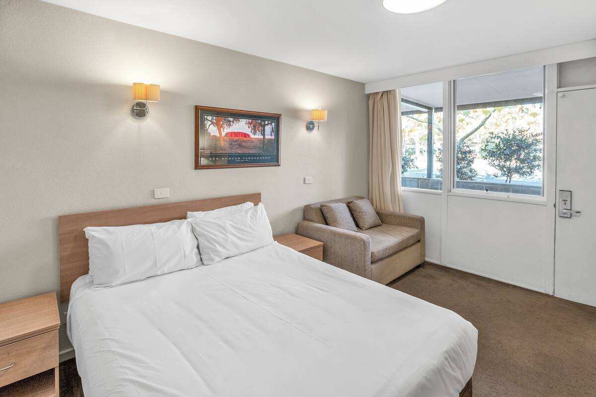 A hotel room with a large bed, a chair, a desk, and a window with curtains.