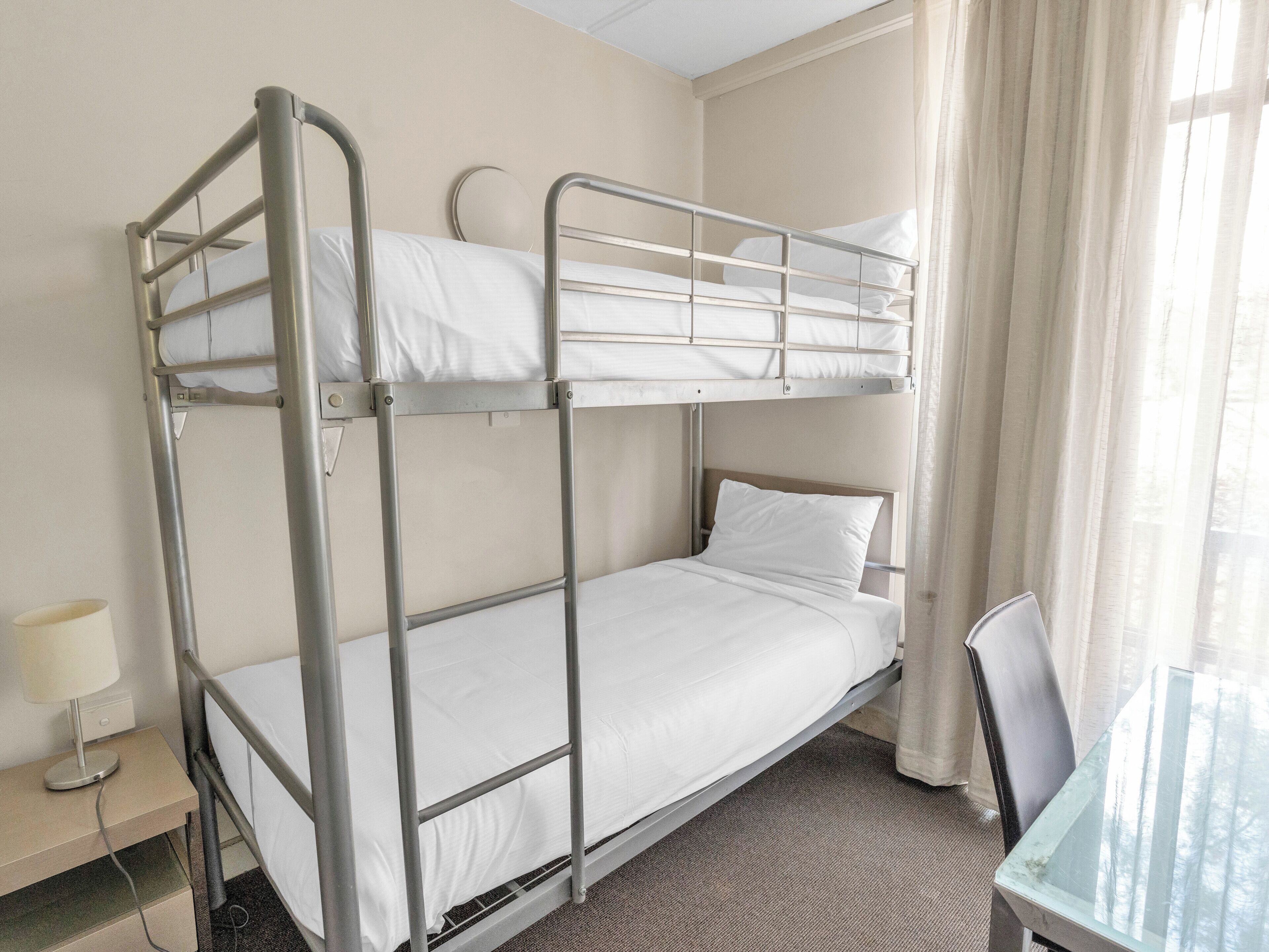 Une chambre avec un lit superposé, un bureau et une chaise, une lampe et une fenêtre avec des rideaux.