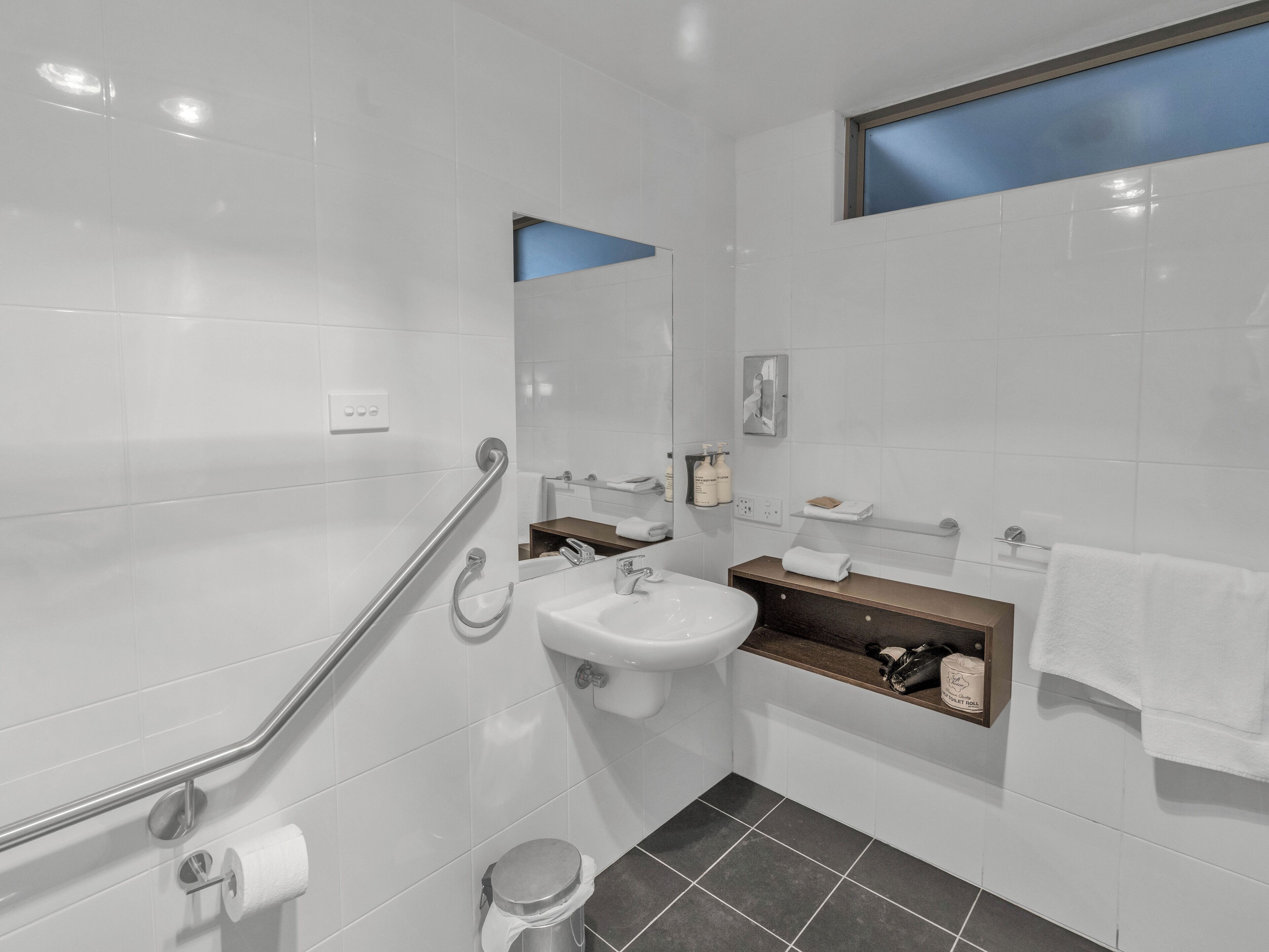 Un baño moderno con baldosas blancas, un lavamanos, un espejo, un perchero para toallas y una ventana.