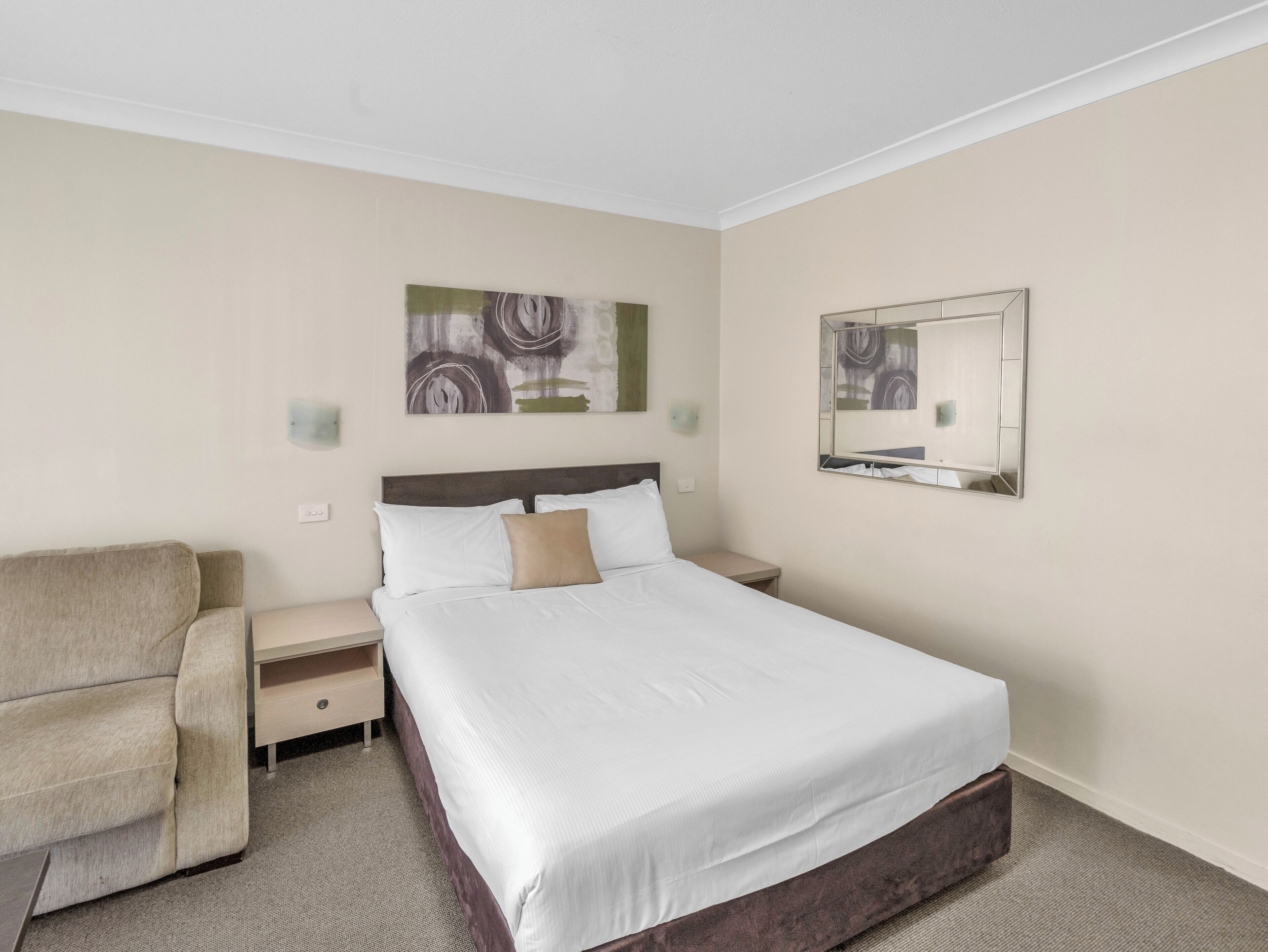 Habitación de hotel con cama, una silla, mesita de noche, espejo montado en la pared y obra de arte abstracta.