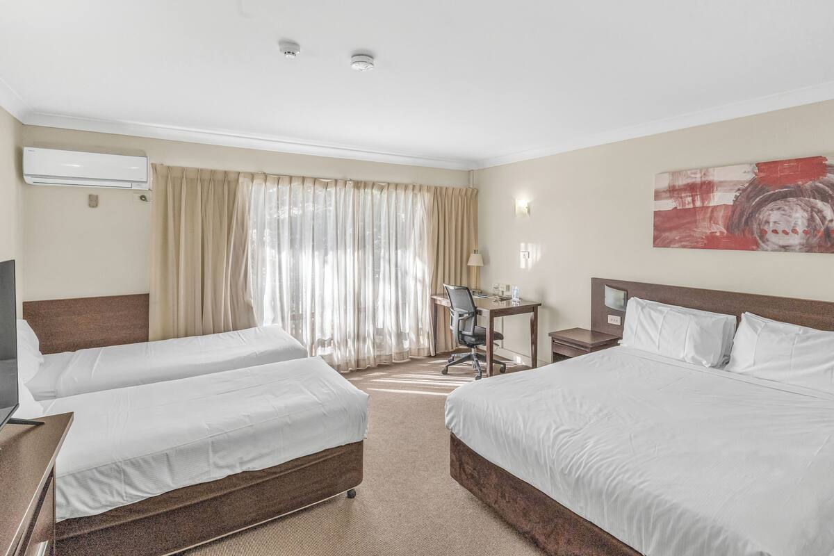 A hotel room with two beds, a desk, a chair, an air conditioning unit, and a large window with curtains.