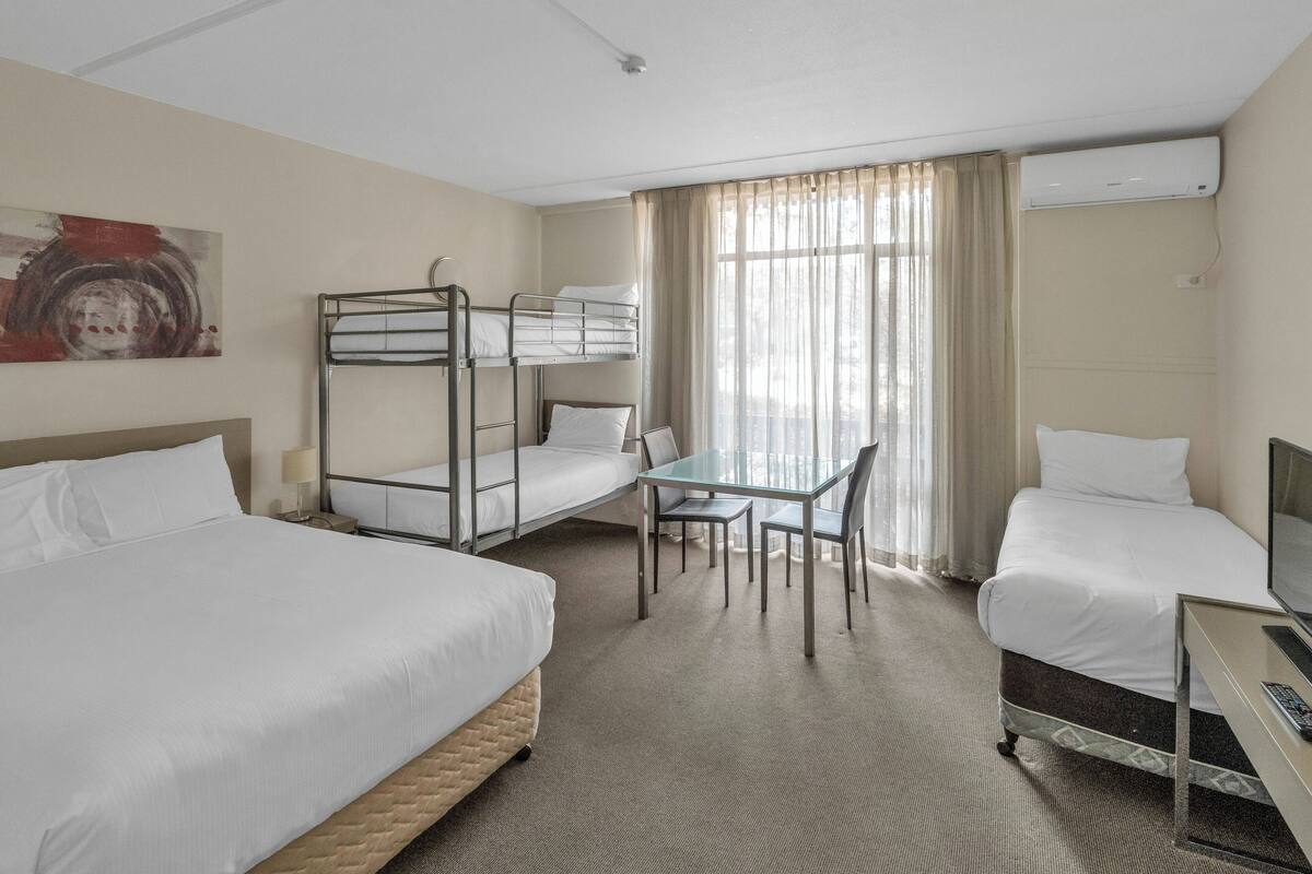 A hotel room with bunk beds, a desk, a chair, a television, and a large window with curtains.