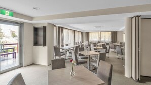 A hotel dining area with multiple tables and chairs, a large window with curtains, and a green emergency exit sign. - Tall Trees Ainslie Motel (Ainslie)
