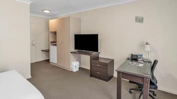 Habitación de hotel con una cama, un escritorio con silla, televisión, kitchenette pequeña y un cubo de basura.