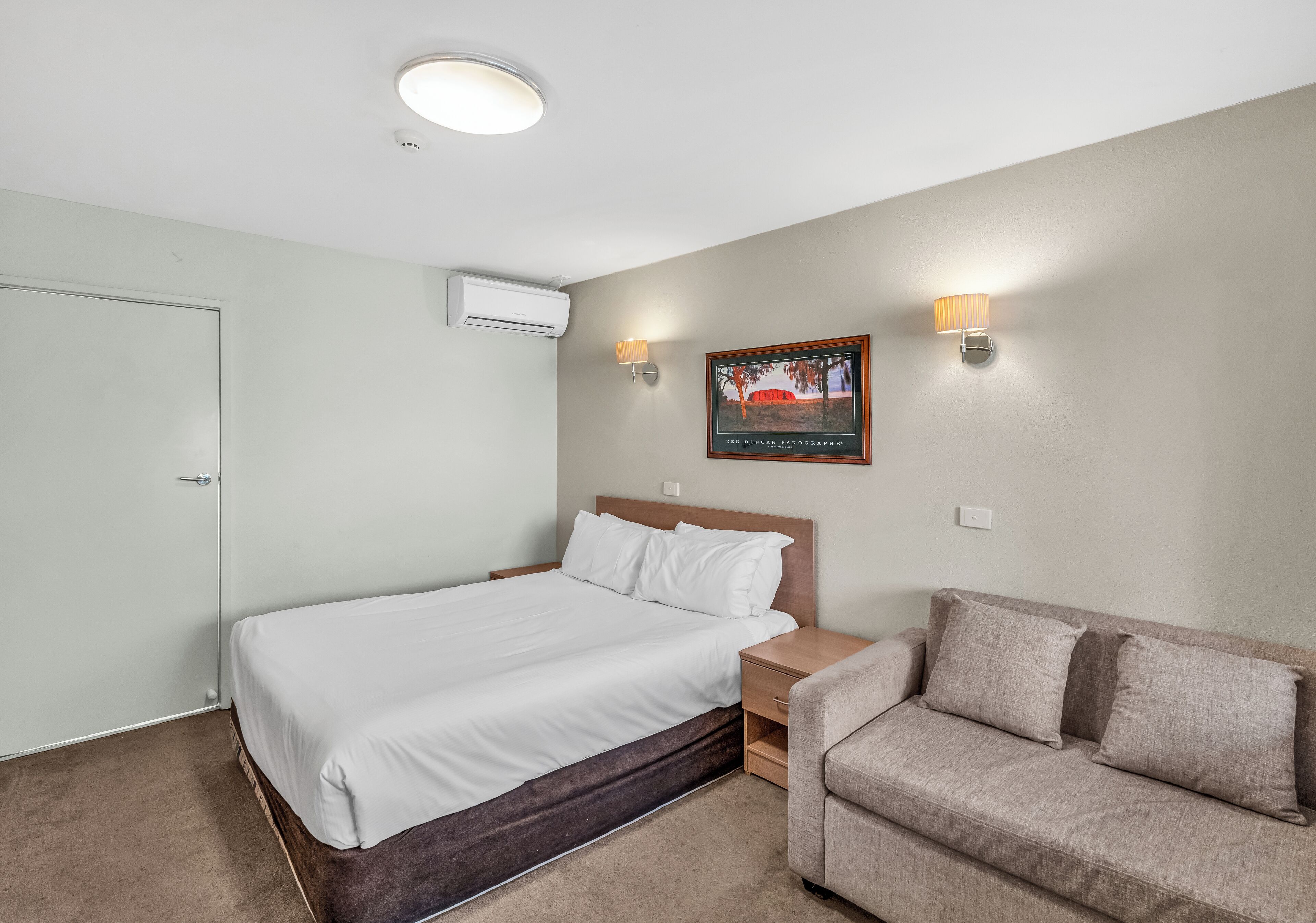 A hotel room with a bed, a nightstand, a lamp, a sofa, and a wall-mounted picture.