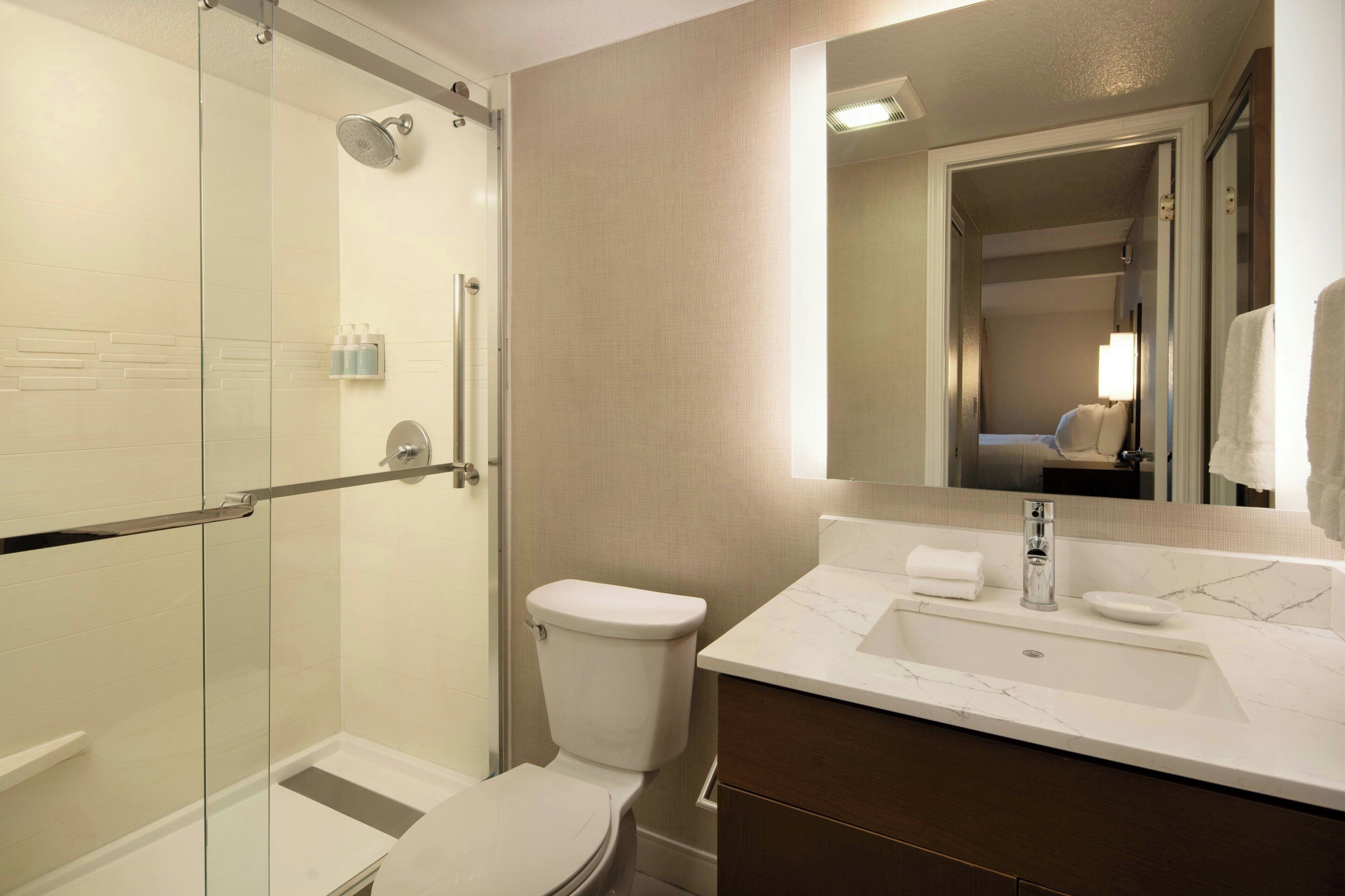 A modern bathroom with a glass-enclosed shower, a white toilet, a marble sink, and a mirror reflecting a room with a bed and a nightstand.