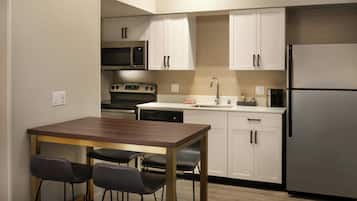 A compact kitchen with white cabinets, a stainless steel refrigerator, a microwave, a dishwasher, and a wooden dining table with four chairs.