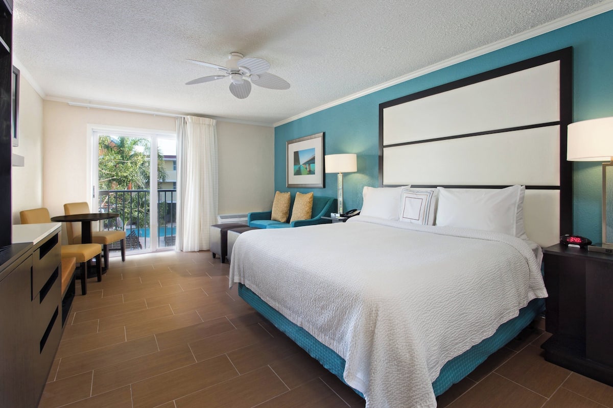 A hotel room with a large bed, a desk, a chair, a balcony with a view, and a ceiling fan.