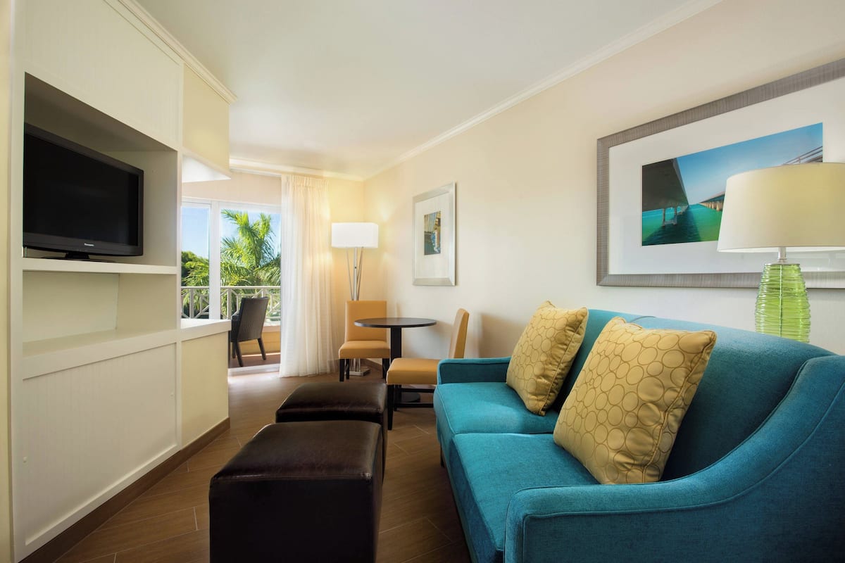 A hotel room with a teal sofa, a flat-screen TV, a dining area with a table and chairs, and a view of palm trees through the window.
