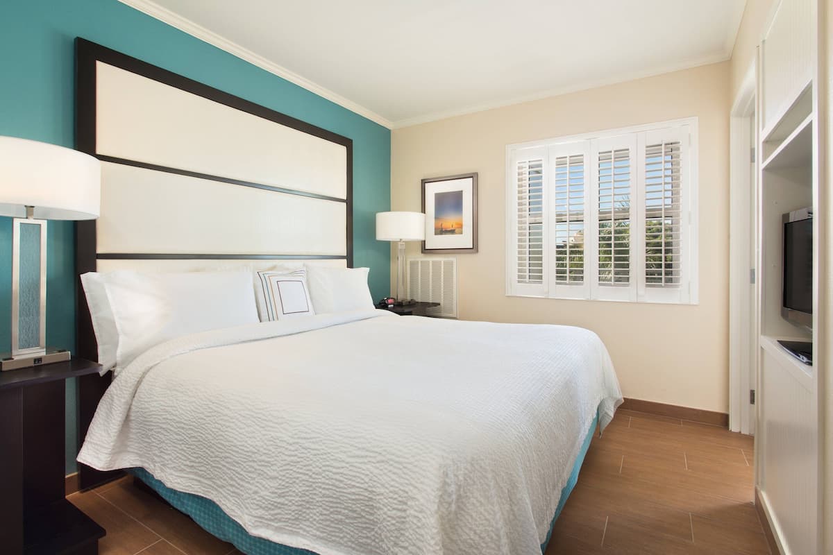 A neatly made bed with white linens, a nightstand with a lamp, a framed picture on the wall, and a window with shutters.
