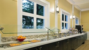 A buffet setup with various food items, a glass display case, and a window with a view of trees. - Fairfield Inn and Suites by Marriott Key West (Key West)