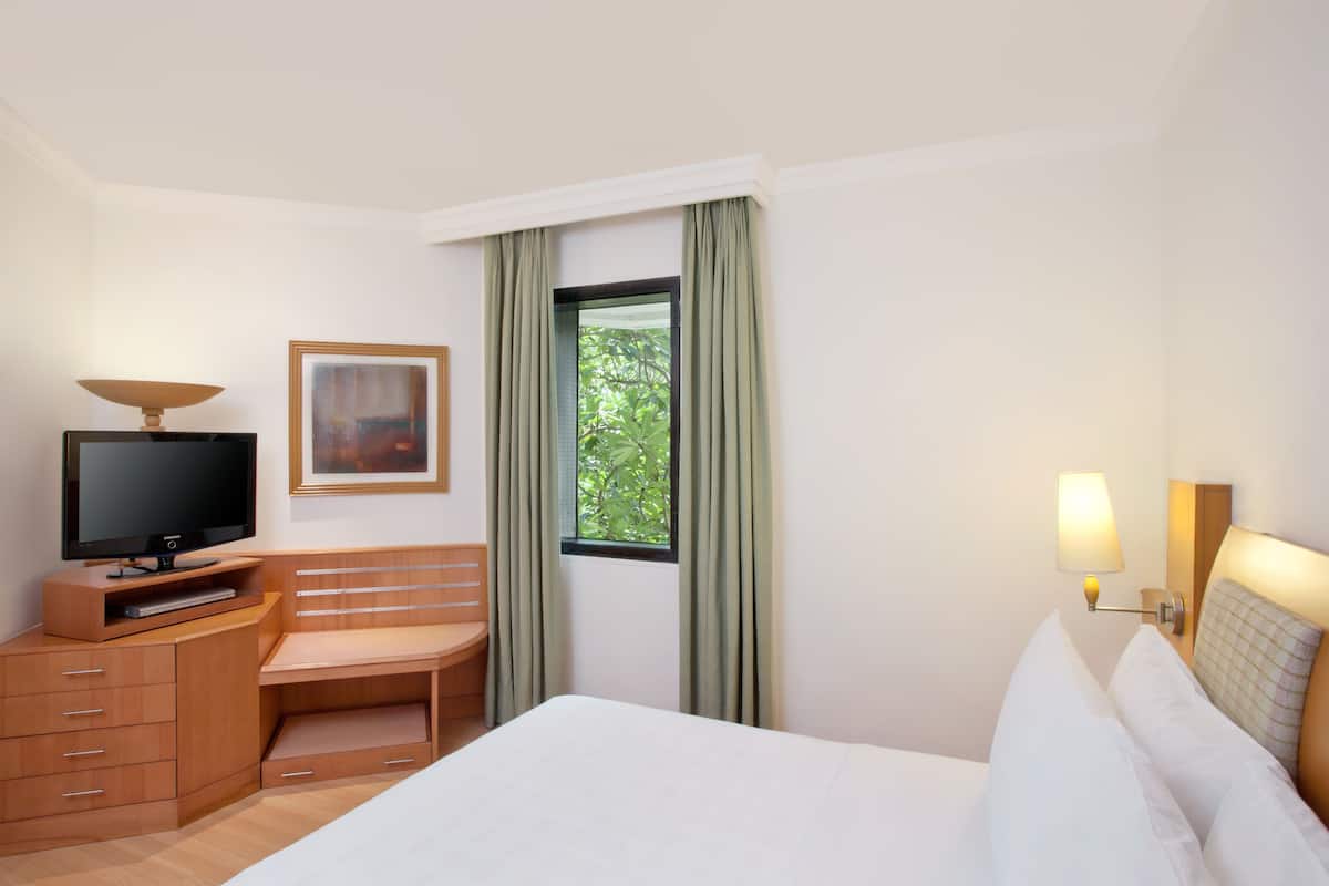 A hotel room with a bed, a desk with a television, a window with curtains, and a wall-mounted lamp.