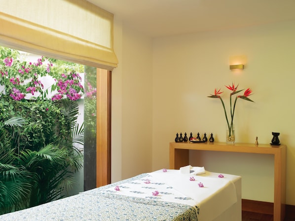 A room with a massage table, a wooden console table with decorative items, and a view of lush greenery and flowers through a window. - Trident, Chennai (Chennai)