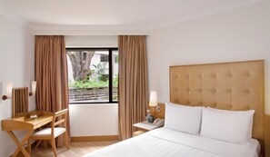 A hotel room with a bed, a desk, a chair, a nightstand, a lamp, and a window with curtains.