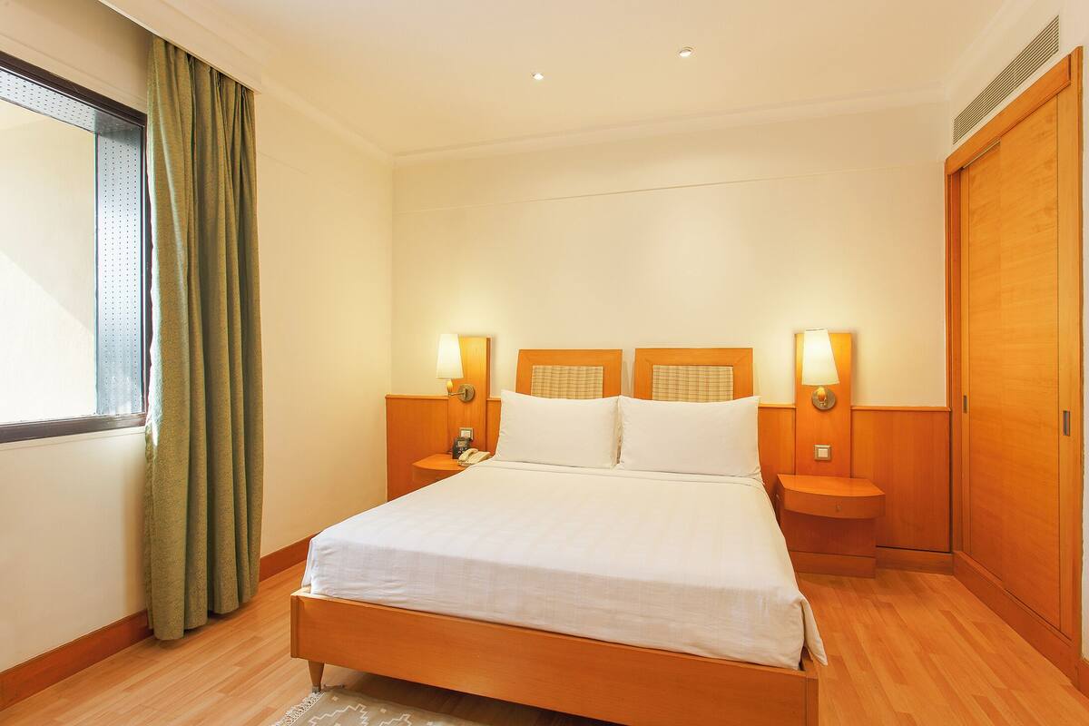 A hotel room with a large bed, wooden headboard, two bedside tables with lamps, a window with curtains, and a wooden wardrobe.