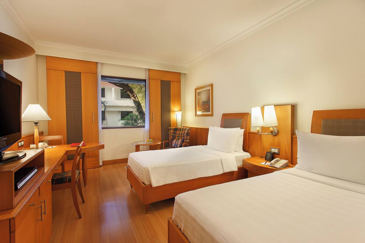 A hotel room with two beds, a desk, a chair, a television, and a window with a view of buildings.