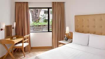 Une chambre d’hôtel comprenant un lit, un bureau, une chaise, deux tables de chevet avec des lampes et une fenêtre avec des rideaux.