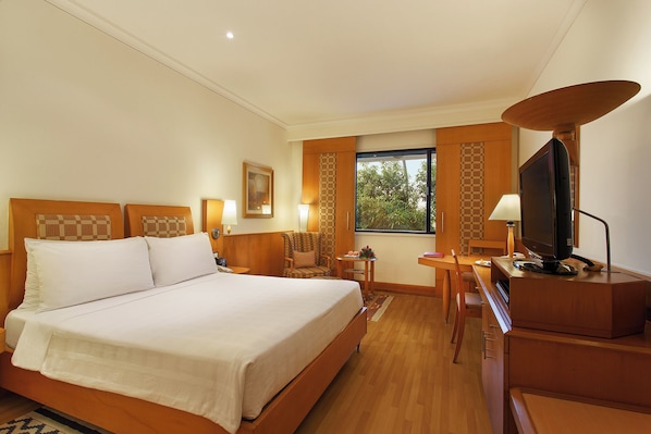 A hotel room with a large bed, a desk with a computer, a television, and a window with a view of trees. - Trident, Chennai (Chennai)