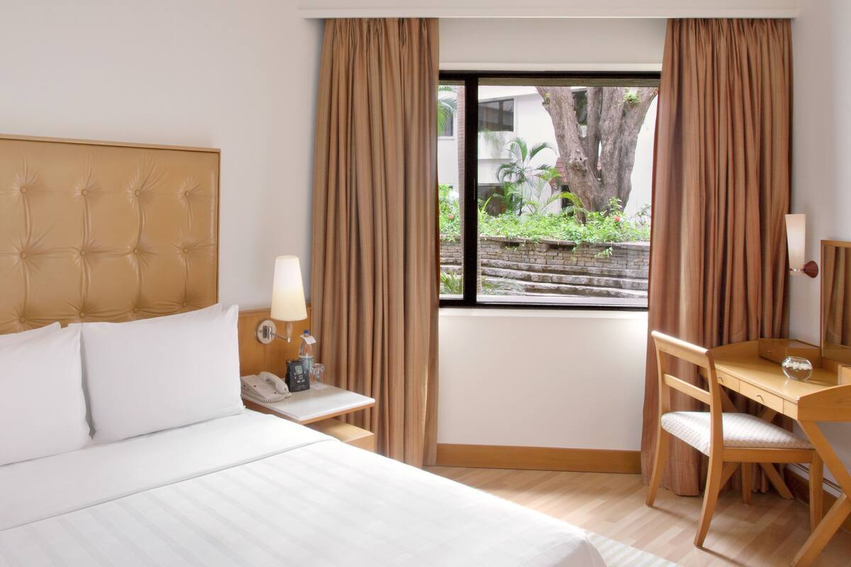 A hotel room with a large bed, a desk, and a chair. There is a window with curtains, a nightstand with a lamp, and a phone.
