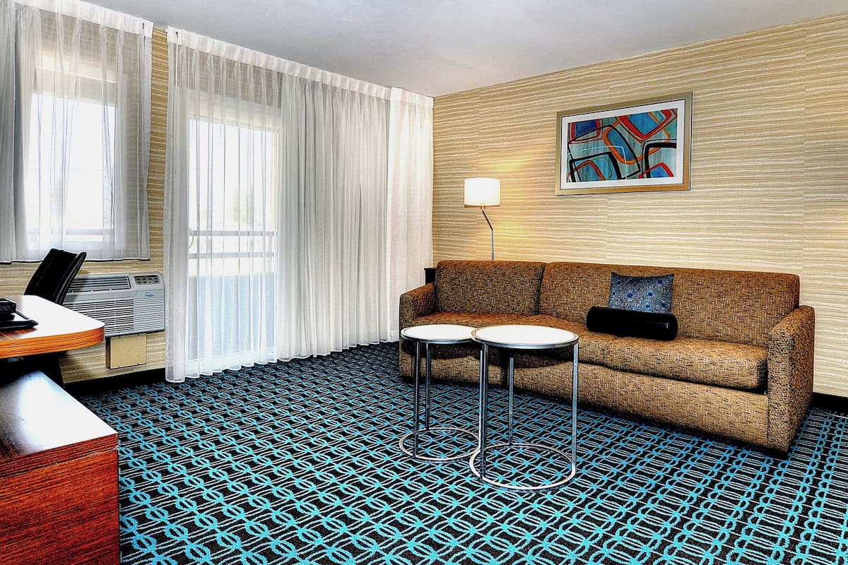 Une chambre d’hôtel avec un canapé, deux petites tables, une lampe, un tableau au mur et un tapis à motifs.