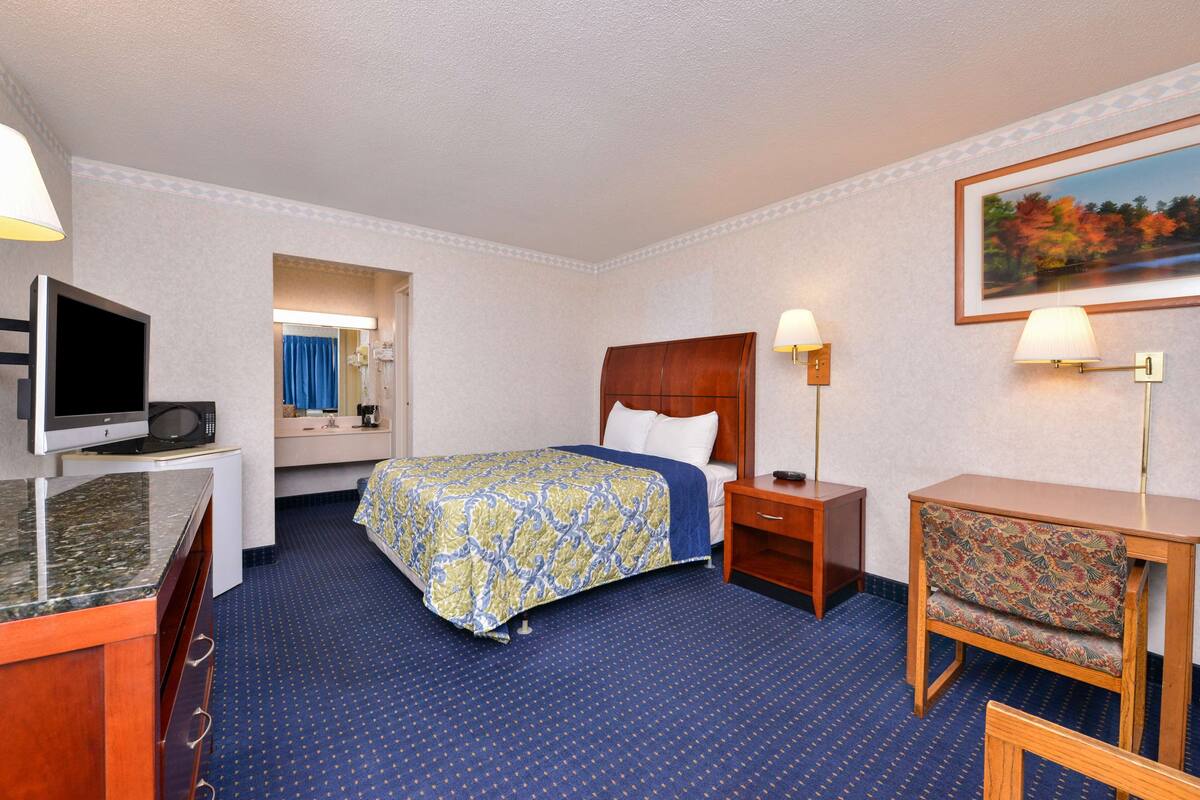 A hotel room with a bed, a desk with a television, a chair, and a painting on the wall.