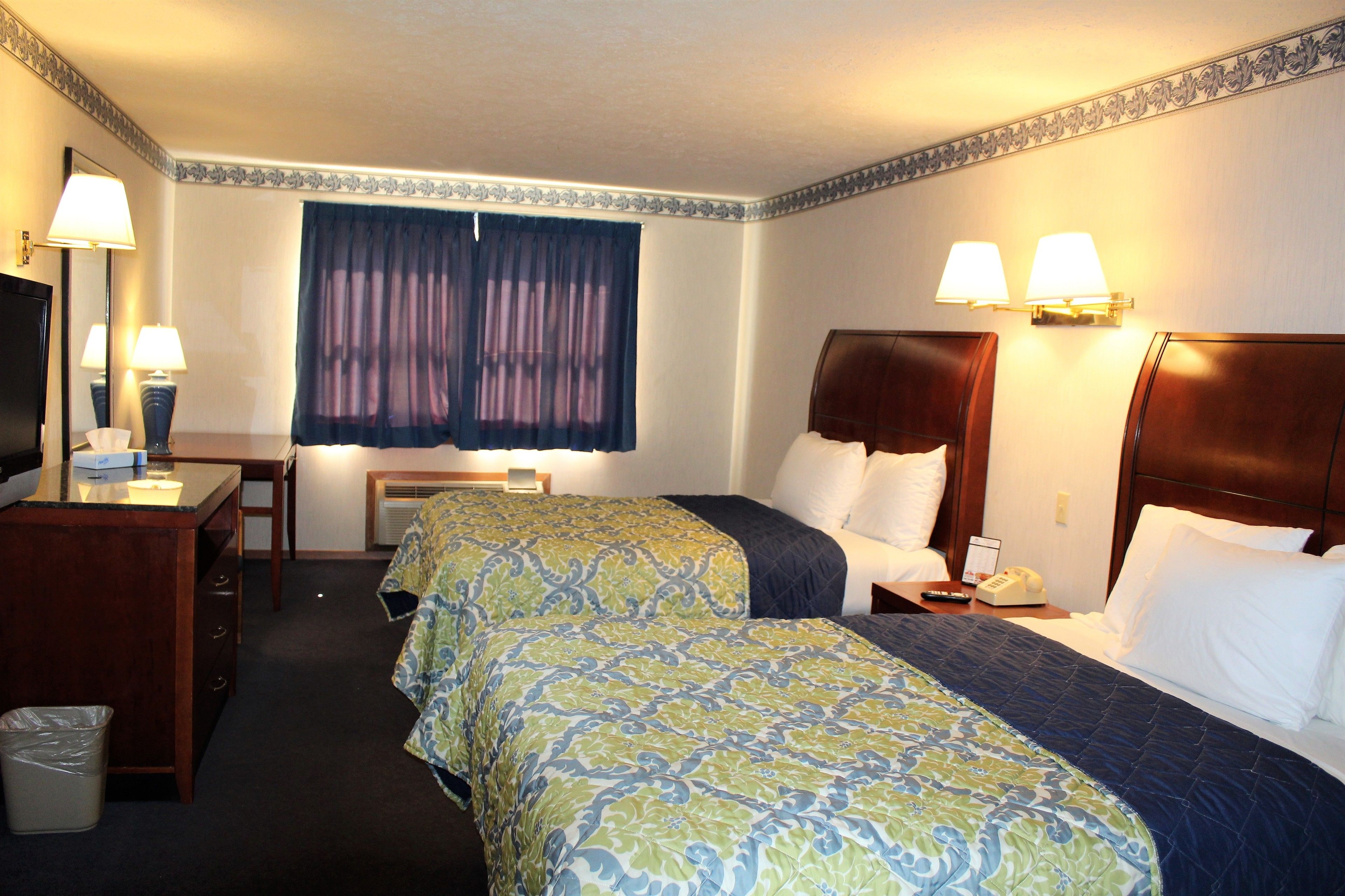 A hotel room with two beds, a desk, a chair, a trash bin, and a window with curtains.