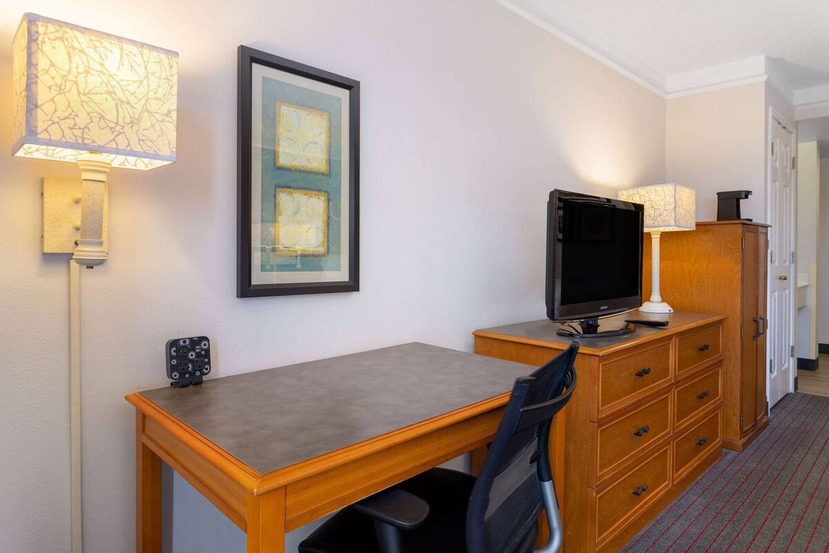 A hotel room with a wooden desk, a television, a chair, a lamp, and a framed picture on the wall.