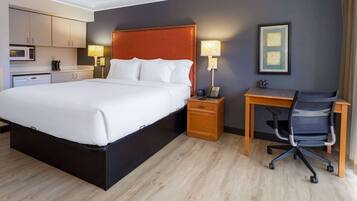 Habitación de hotel con una cama grande, un escritorio con silla, una mesita de noche, una lámpara, un cuadro en la pared y una kitchenette con microondas.