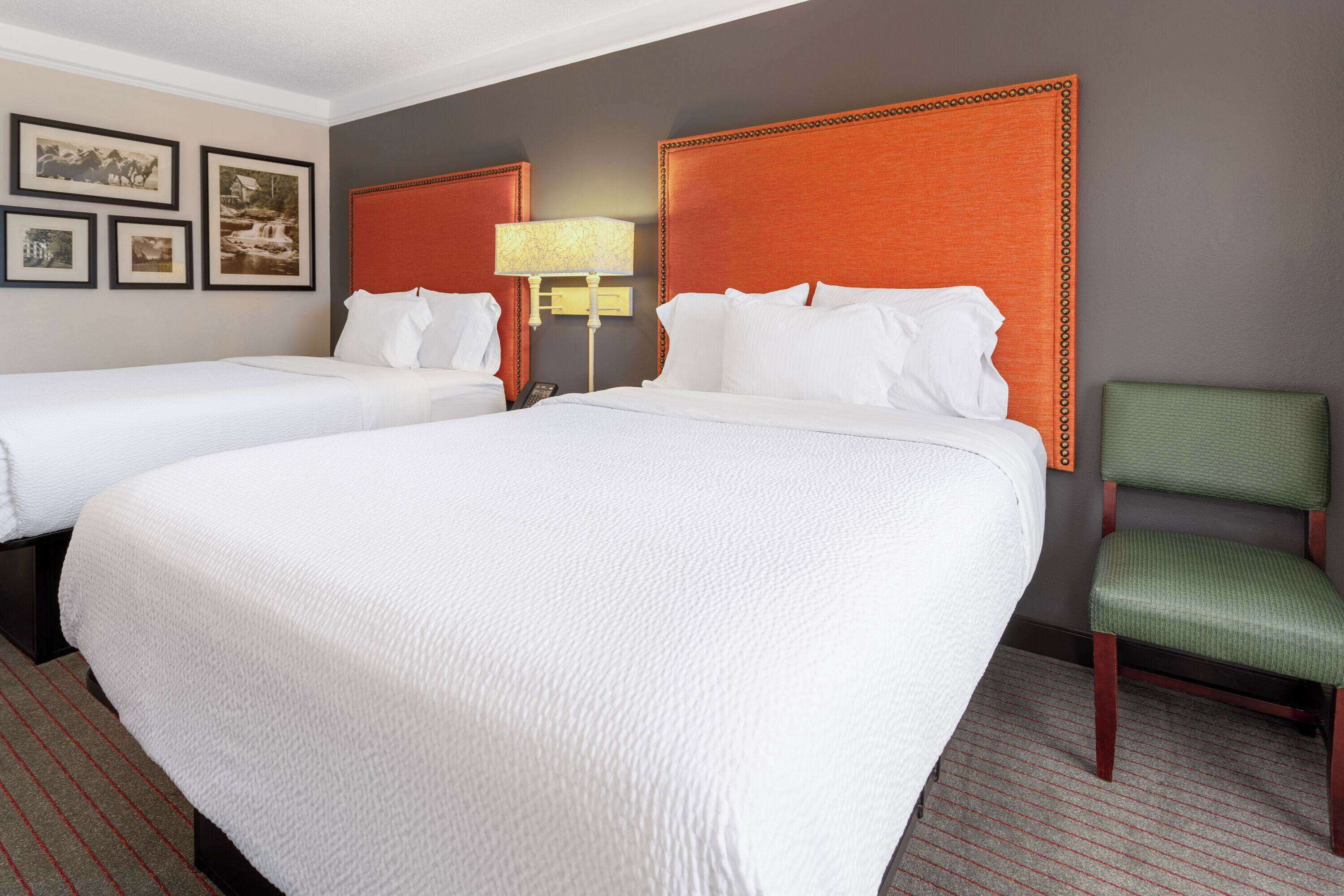 A hotel room with two beds, a green chair, and a lamp.