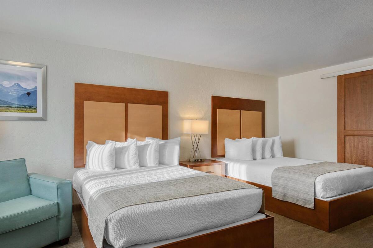 A hotel room with two beds, a nightstand with a lamp, a framed picture on the wall, and a light-colored armchair.