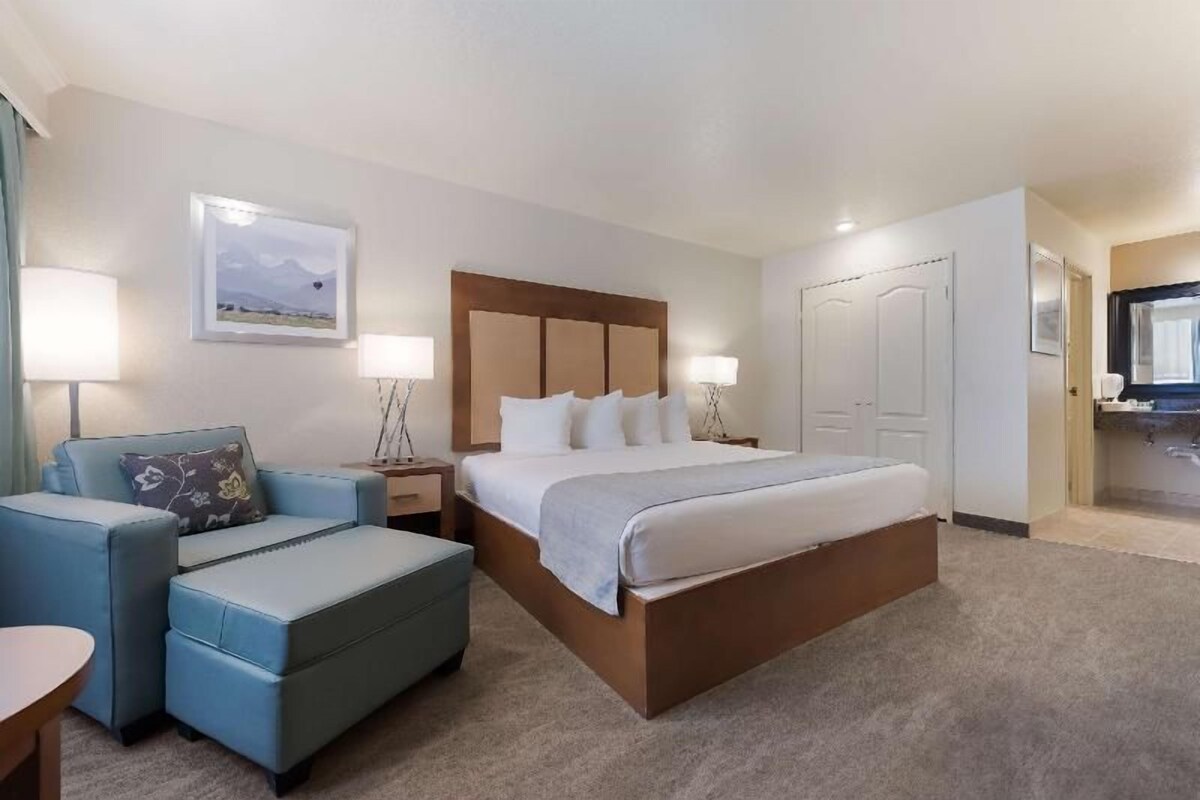 A hotel room with a large bed, a seating area with a chair and ottoman, a desk, and a bathroom visible through an open door.