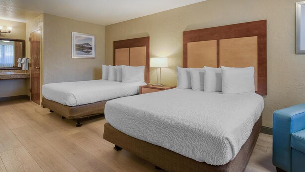 A hotel room with two beds, a wooden headboard, a bedside table with a lamp, a framed picture on the wall, and a bathroom with a mirror and towel rack.