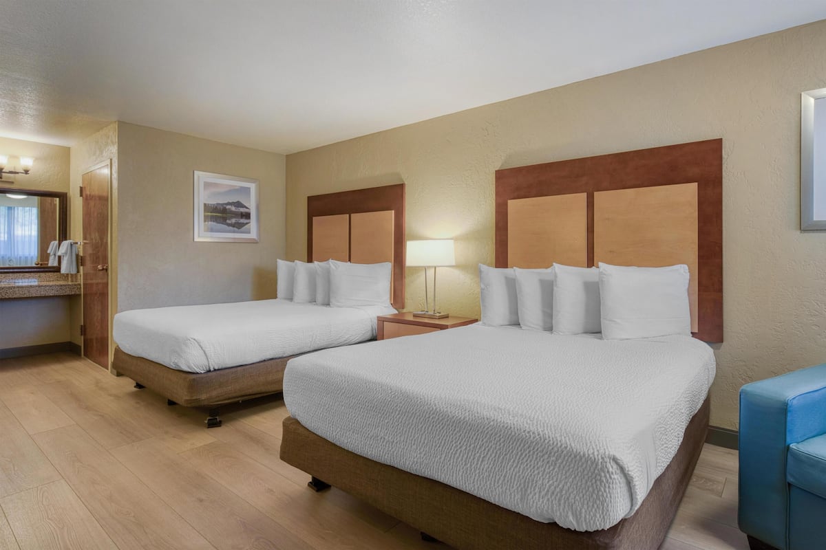 A hotel room with two beds, a wooden headboard, a bedside table with a lamp, a framed picture on the wall, and a bathroom with a mirror and towel rack.
