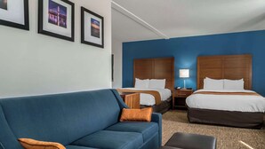 A hotel room with a blue sofa, two beds with wooden headboards, a small table, and a lamp.