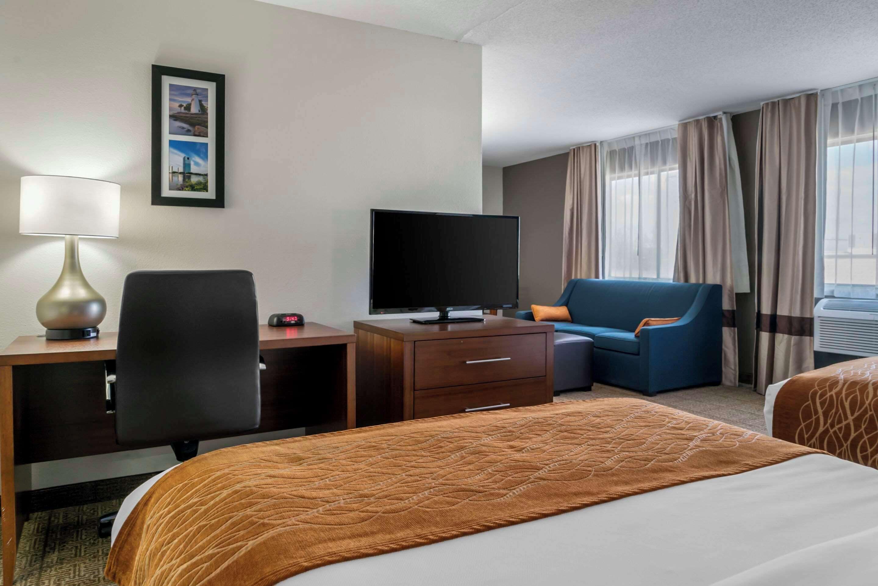 A hotel room with a bed, a desk with a lamp, a television, a chair, and a window with curtains.