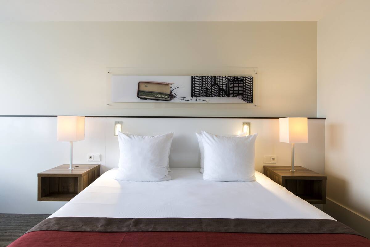 A hotel room with a large bed, two bedside tables, a wall-mounted artwork, and a red accent on the bedspread.