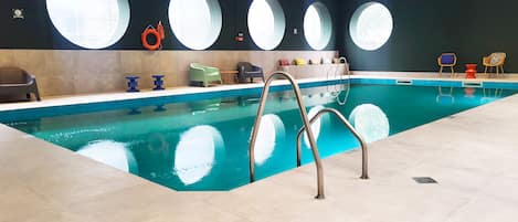 An indoor swimming pool with a clear view of the ceiling, a ladder, and a row of chairs along the poolside.