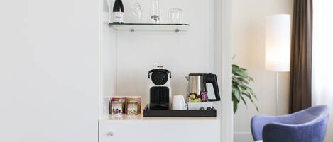 A hotel room with a minibar, a coffee maker, a wine bottle, and a glass shelf.