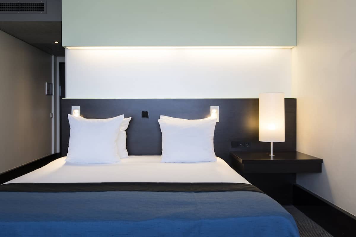 A modern hotel room with a large bed, two white pillows, a bedside table with a lamp, and a headboard with integrated lighting.