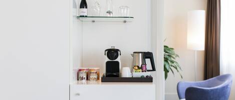 A hotel room with a minibar, a coffee maker, a kettle, and a glass shelf with a bottle and glasses.