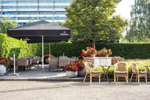 An outdoor seating area with wicker chairs and tables, a Heineken branded umbrella, and potted plants with red flowers.
