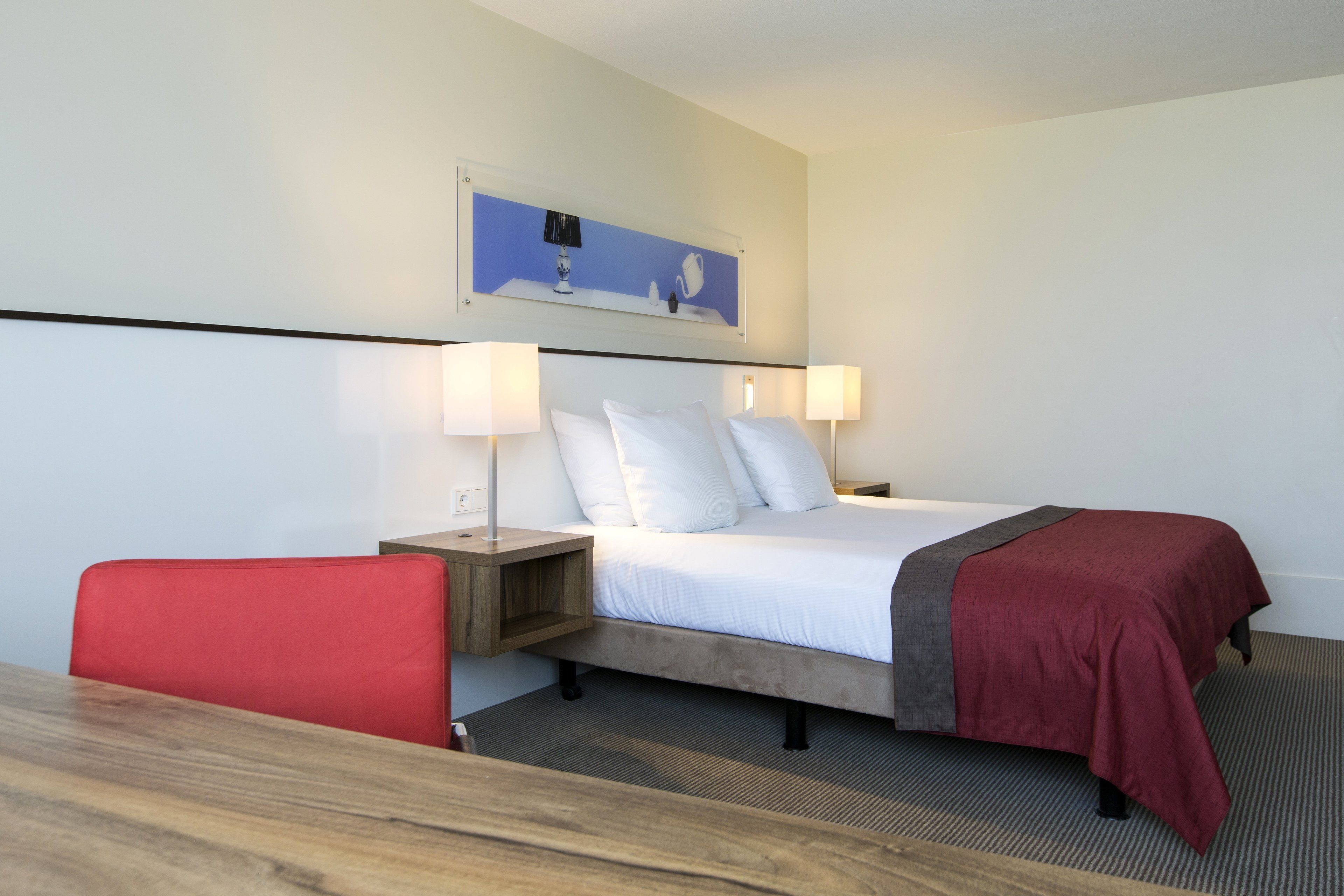 A hotel room with a bed, two bedside lamps, a red chair, a wooden nightstand, and a framed picture on the wall.