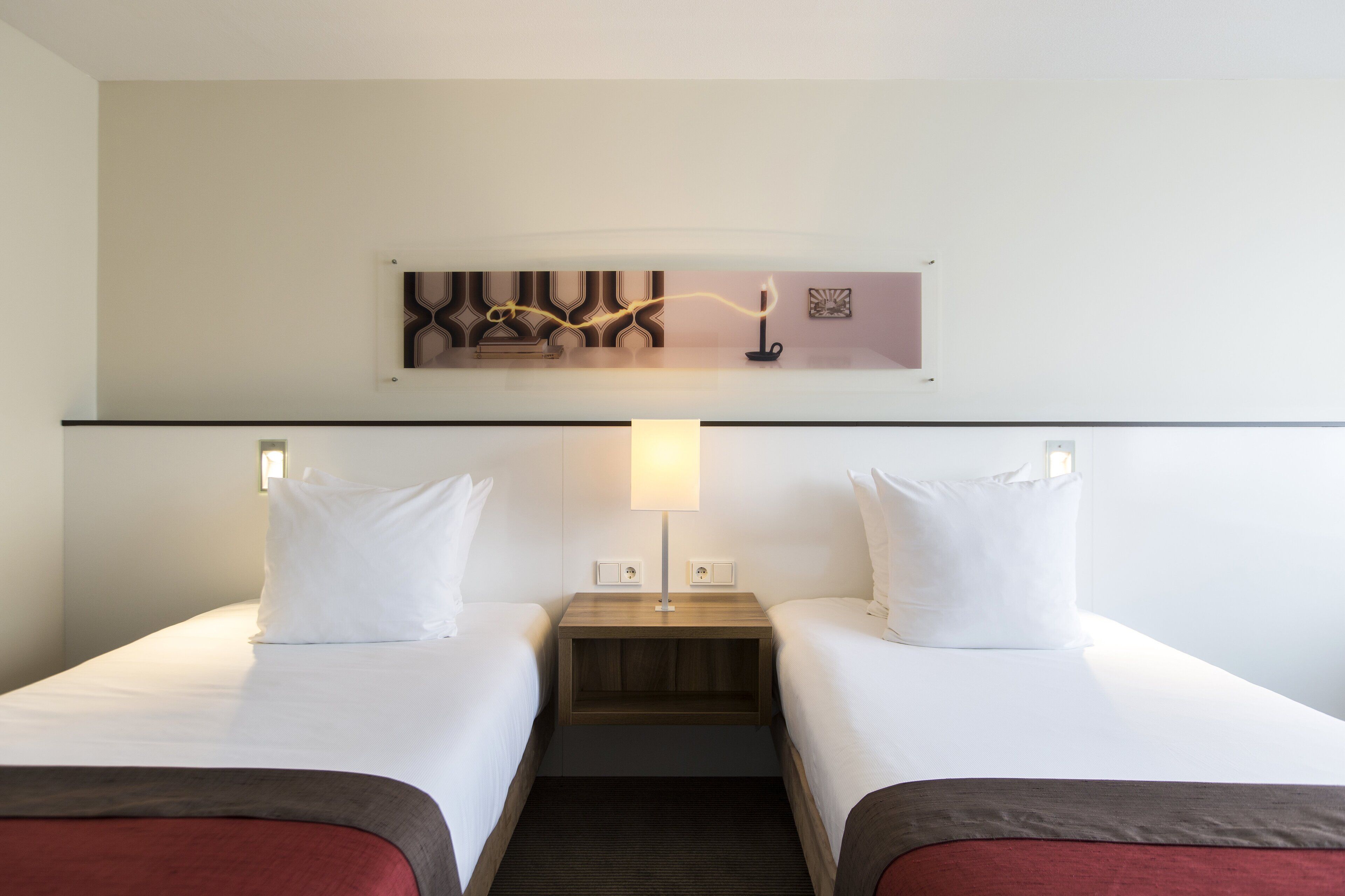 Habitación de hotel con dos camas individuales, una mesita de noche con lámpara y una obra de arte colgada en la pared.