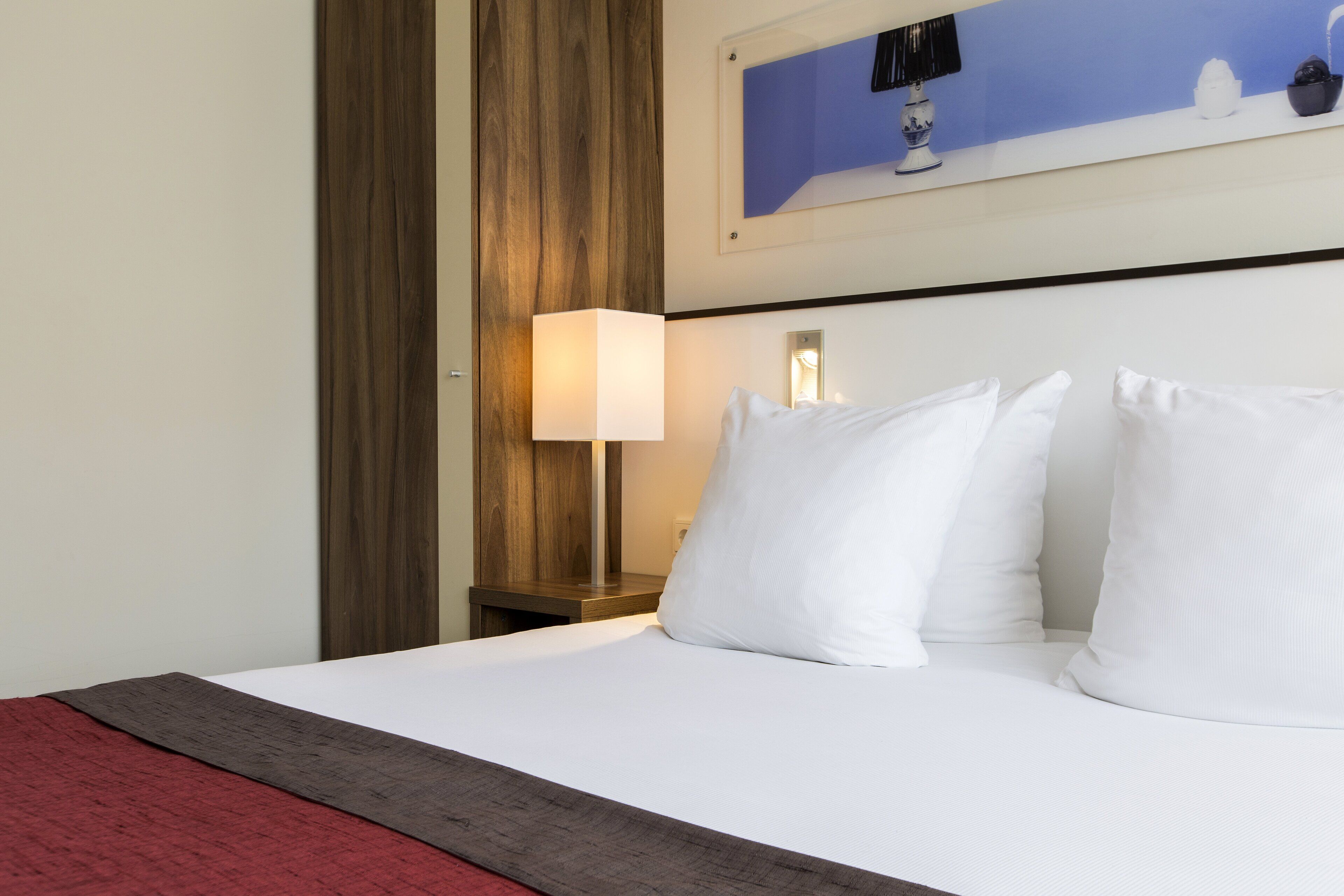 Une chambre d’hôtel avec un lit bien fait, une table de chevet en bois avec une lampe et un tableau accroché au mur.