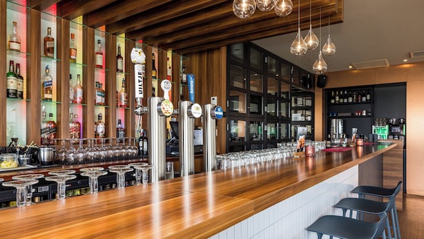 Una zona de bar con una barra de madera larga, múltiples grifos y una variedad de botellas y vasos.