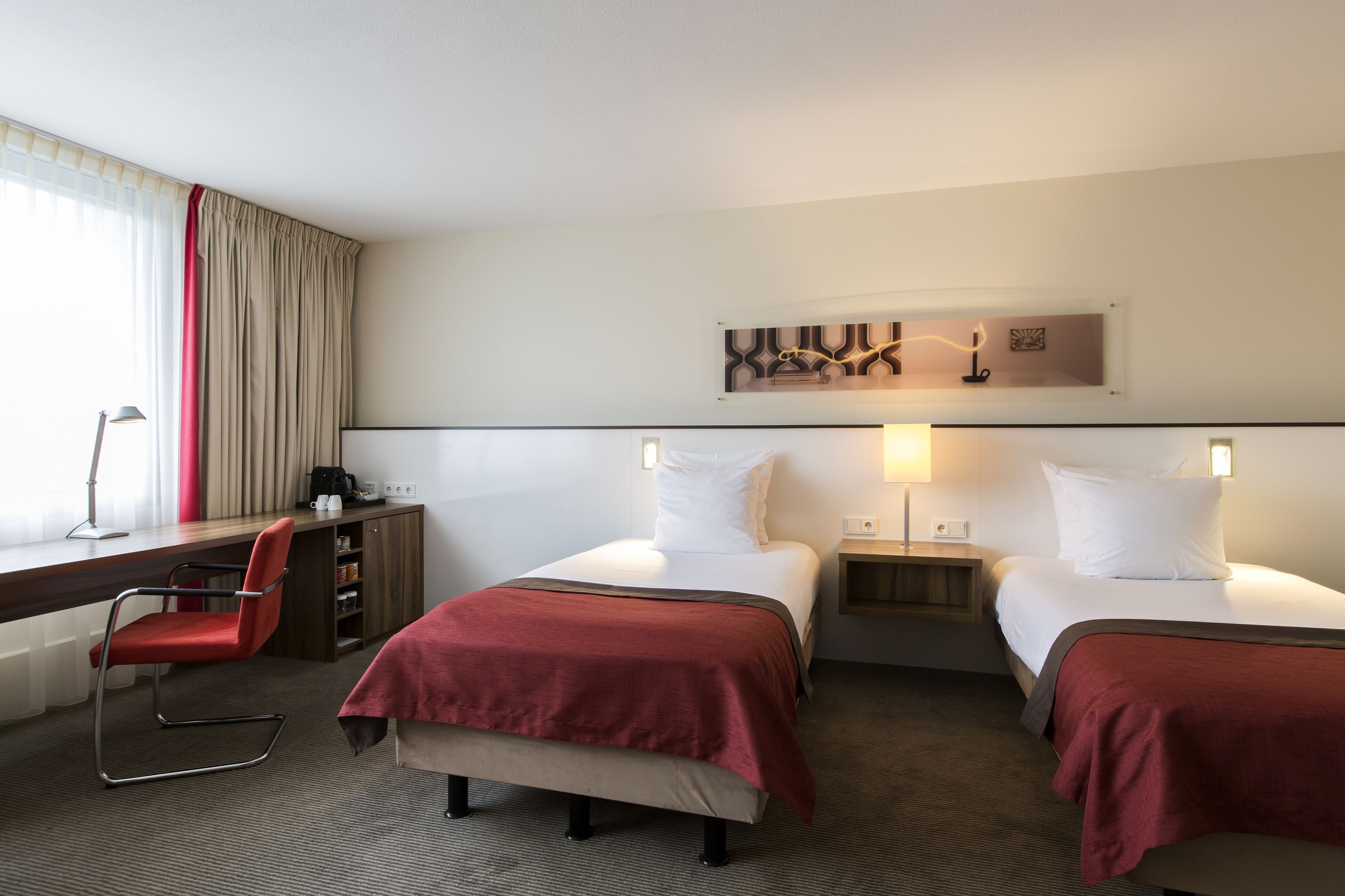 Ein Hotelzimmer mit zwei Betten, einem Schreibtisch, einem Stuhl und einem Fenster mit Vorhängen.