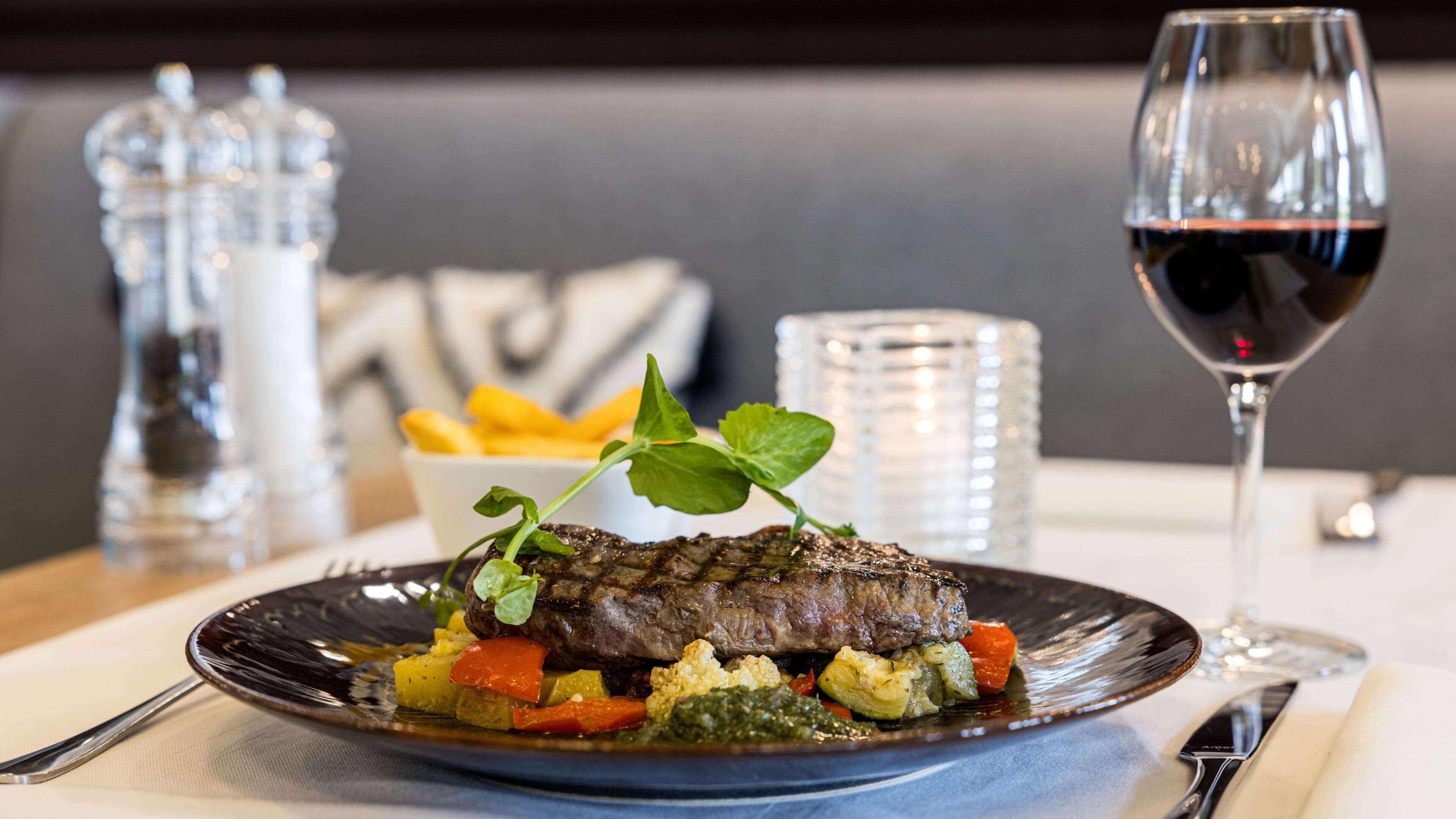 Un plato de carne con verduras y una copa de vino tinto sobre una mesa.