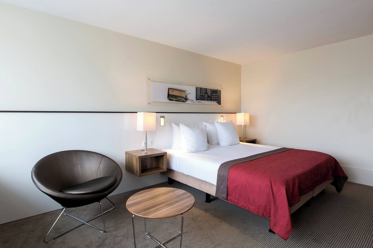 A hotel room with a bed, a chair, a small table, and bedside lamps.